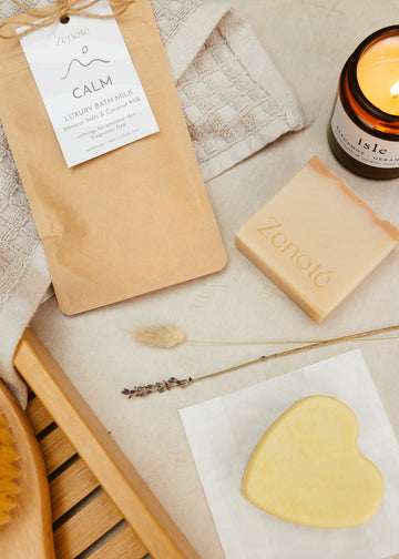 Kraft Compostable Bag with small white label reading Calm Luxury Bath Milk with a square white soap with pink topping. A heart shaped cream moisturiser bar, positioned next to a glowing candle, a bath towel and accessories.