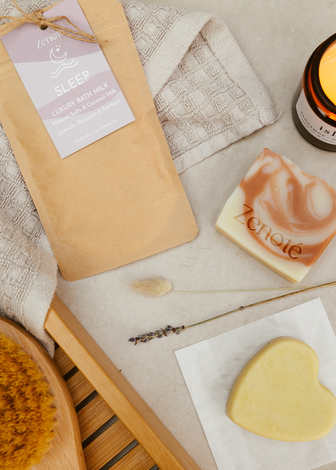 Kraft Compostable Bag with a pink label reading , Sleep Luxury Bath Milk with a square white  & pink swirled soap. A heart shaped cream moisturiser bar, positioned next to a glowing candle, a  bath towel and accessories. 