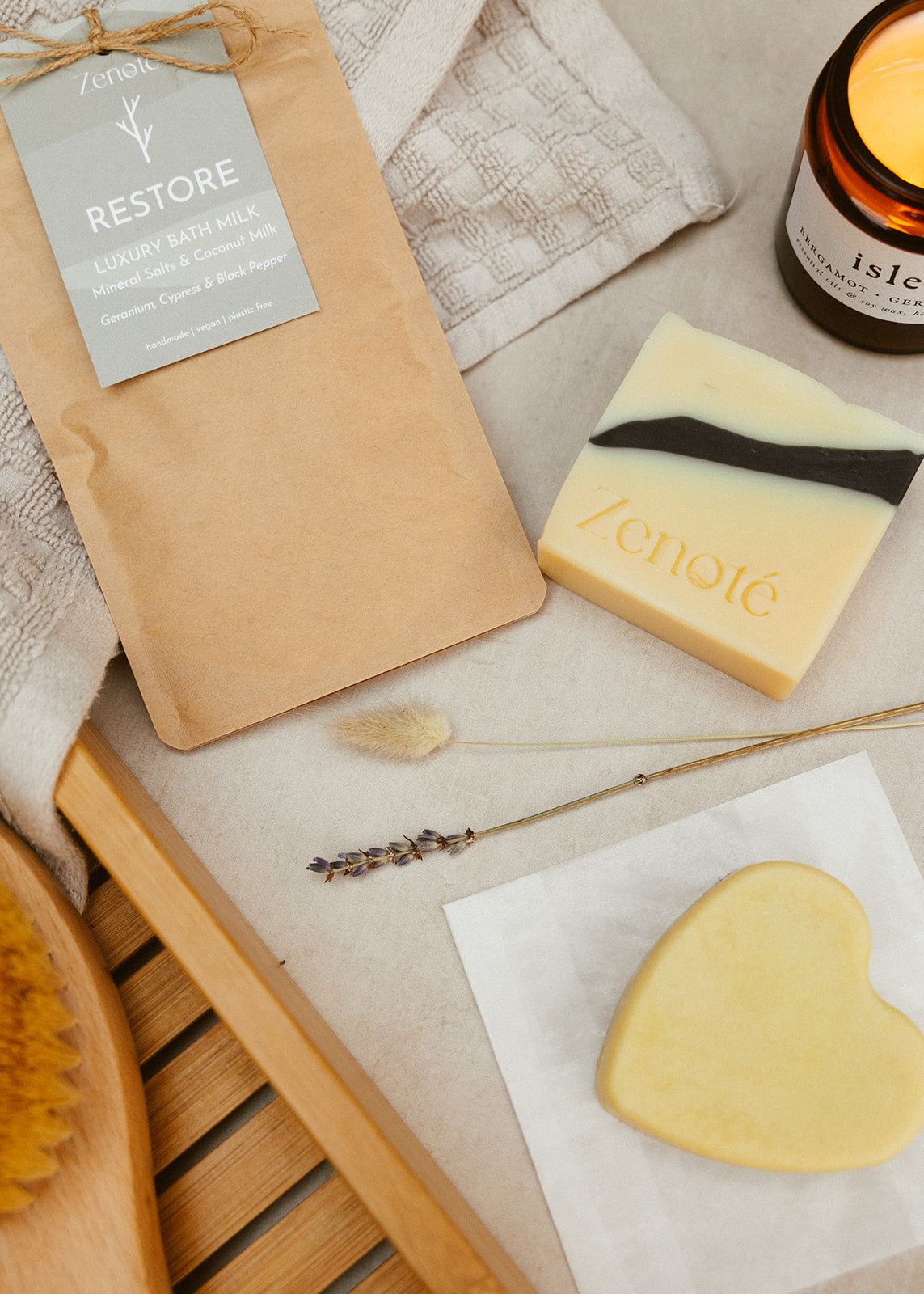 A Compostable Brown Bag with small Green tag  reading Restore Luxury Bath Milk with a square yellow soap with a black stripe running across it. A heart shaped cream moisturiser bar, positioned next to a glowing candle, a  bath towel and accessories. 